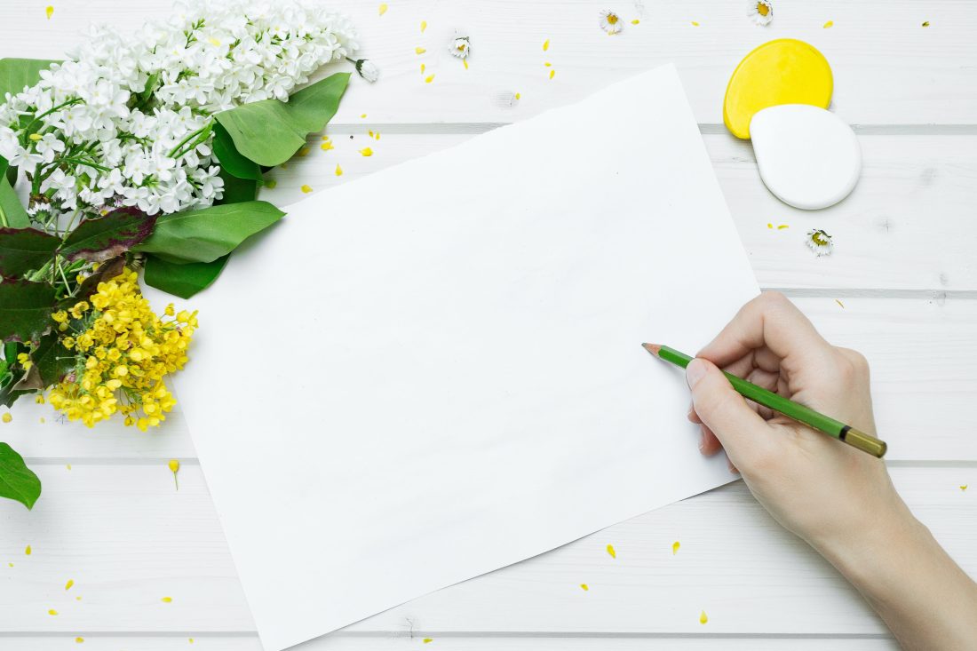 woman's hand holding a pen over white paper next to a bouquet of yellow and white flowers