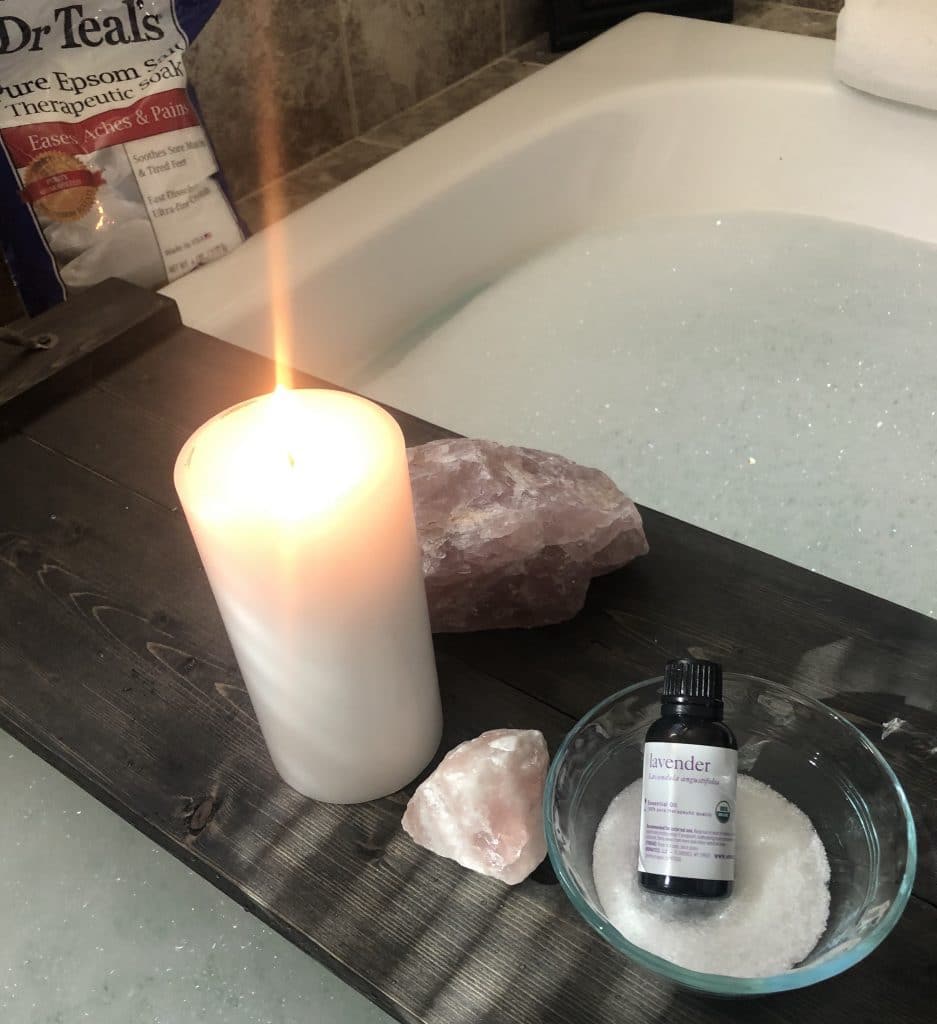 tray atop a filled bath tub holding a candle, rose quartz, epsom salts, and bottle of lavender