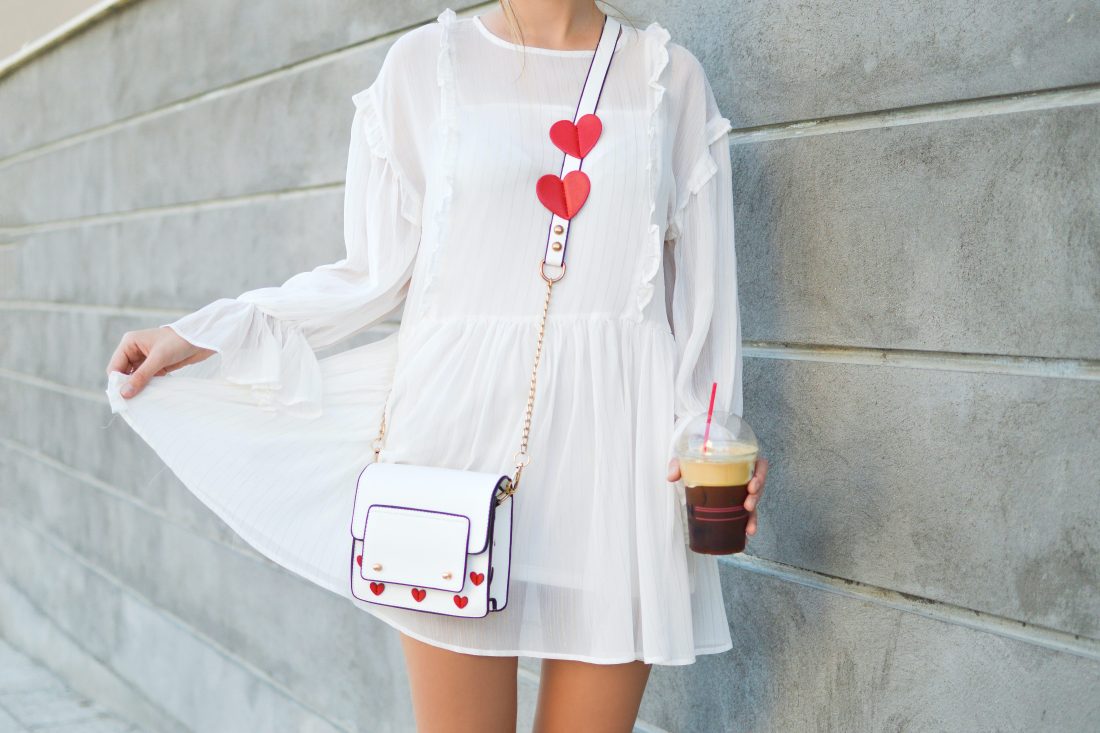 woman from the neck down wearing a white dress and white purse with read hearts holding an iced coffee