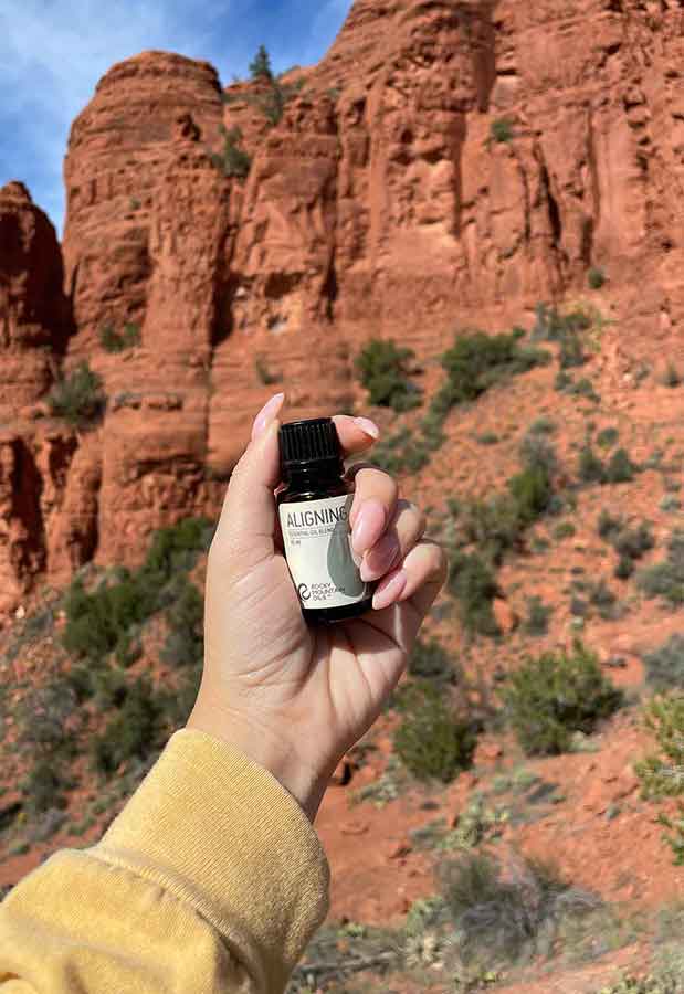 woman's hand holding a bottle of RMO's Aligning essential oil blend with a mountain in the background