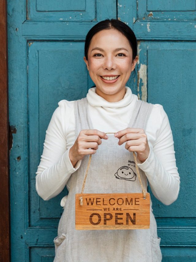 woman standing in front of blue door holding 'welcome, we're open' sign.