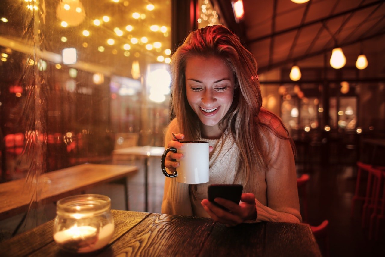 smiling woman holding ceramic mug looking at her cell phone at night